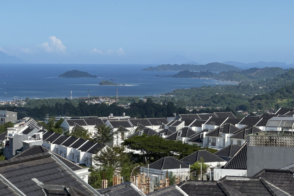 Pemandangan kawasan rumah dekat pantai di area perbukitan dengan deretan rumah beratap gelap di bagian depan, menghadap ke laut biru luas dengan pulau-pulau kecil di kejauhan. Foto diambil dari sudut tinggi pada siang hari cerah, menampilkan perpaduan area hunian, pepohonan hijau, garis pantai, dan panorama laut yang memberi kesan premium dan dekat dengan alam.