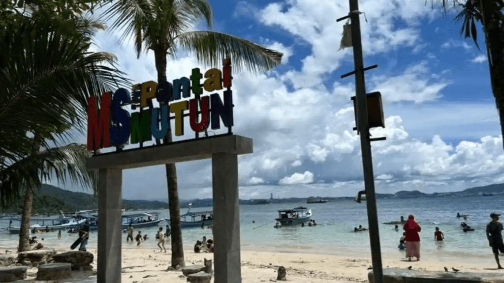 Tropical beach with a colorful sign overhead, palm trees, and people swimming near the shore.