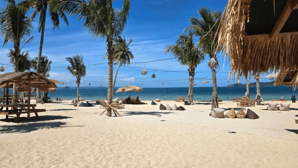 Sunny tropical beach with palm trees, string lights, and wooden huts overlooking turquoise water and white sand.
