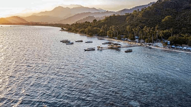 Sunset over a calm bay with boats near a wooden pier and a palm-lined beach by forested hills in the distance, dusk light in the sky.
