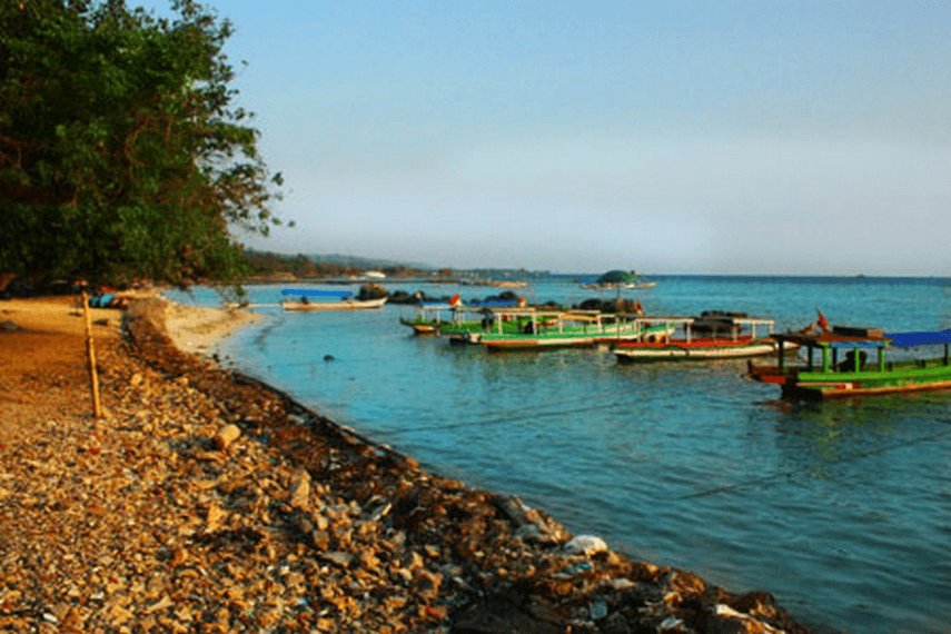 Pebble beach with calm turquoise water and several colorful boats anchored near the shore.