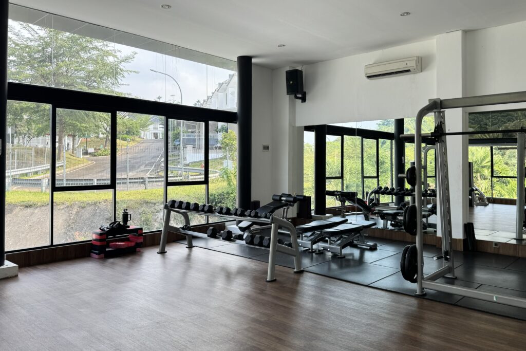 Bright gym with large windows, free weights on racks and a mirror wall, black and gray equipment scattered around the room.